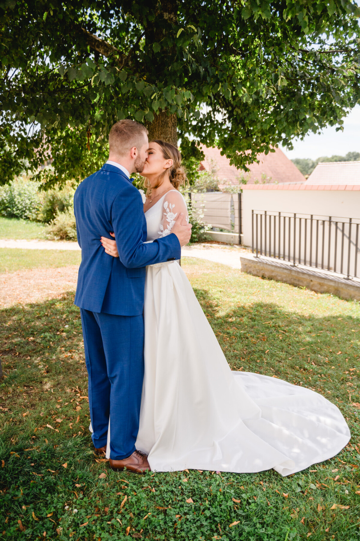 Eine Braut im weißen Kleid und ein Bräutigam im blauen Anzug küssen sich an einem sonnigen Tag unter einem Baum.