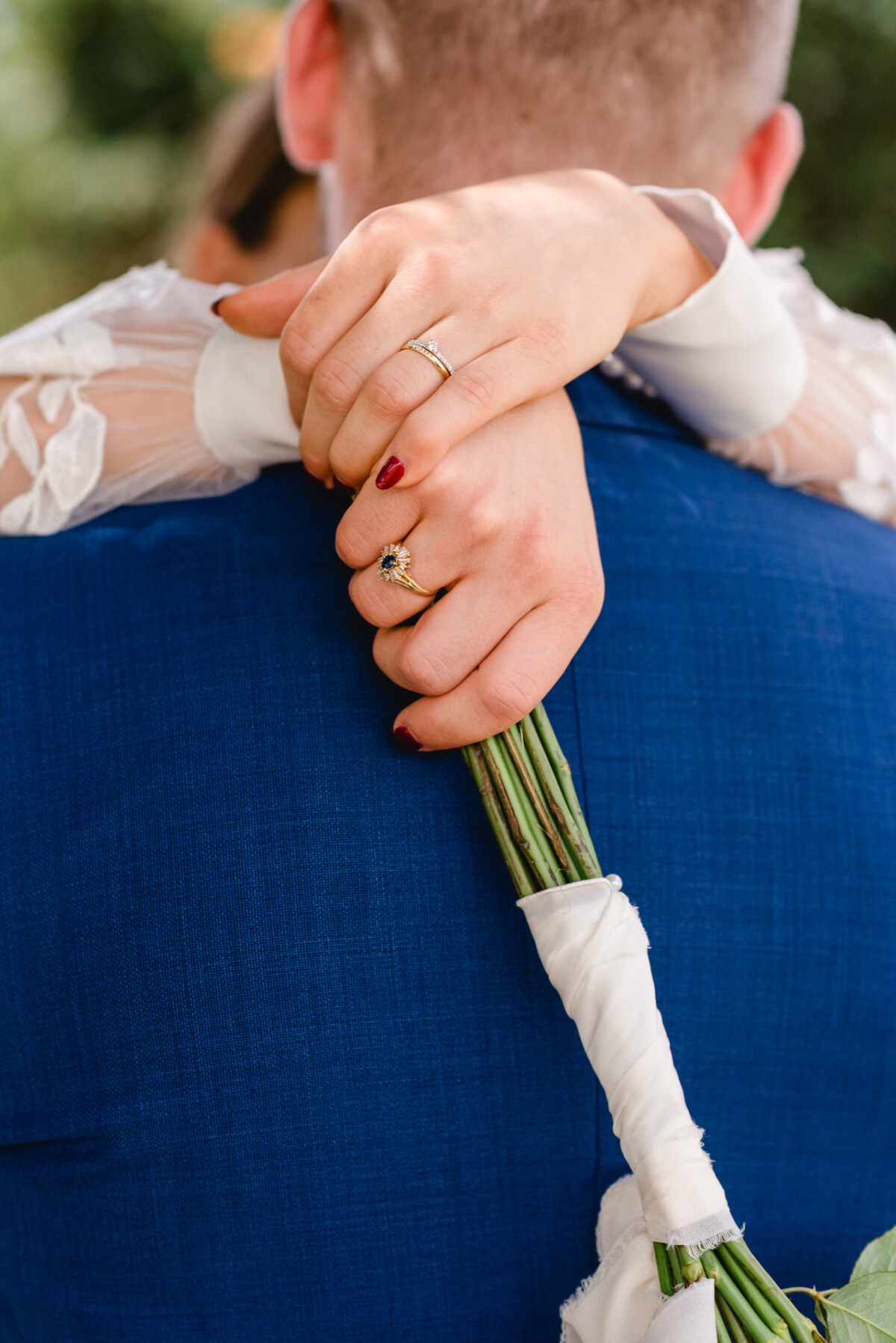 Nahaufnahme einer Person, die eine andere von hinten umarmt. Sie trägt Ringe und hält vor einem blauen Anzug einen mit einem weißen Band umwickelten Blumenstrauß in der Hand.