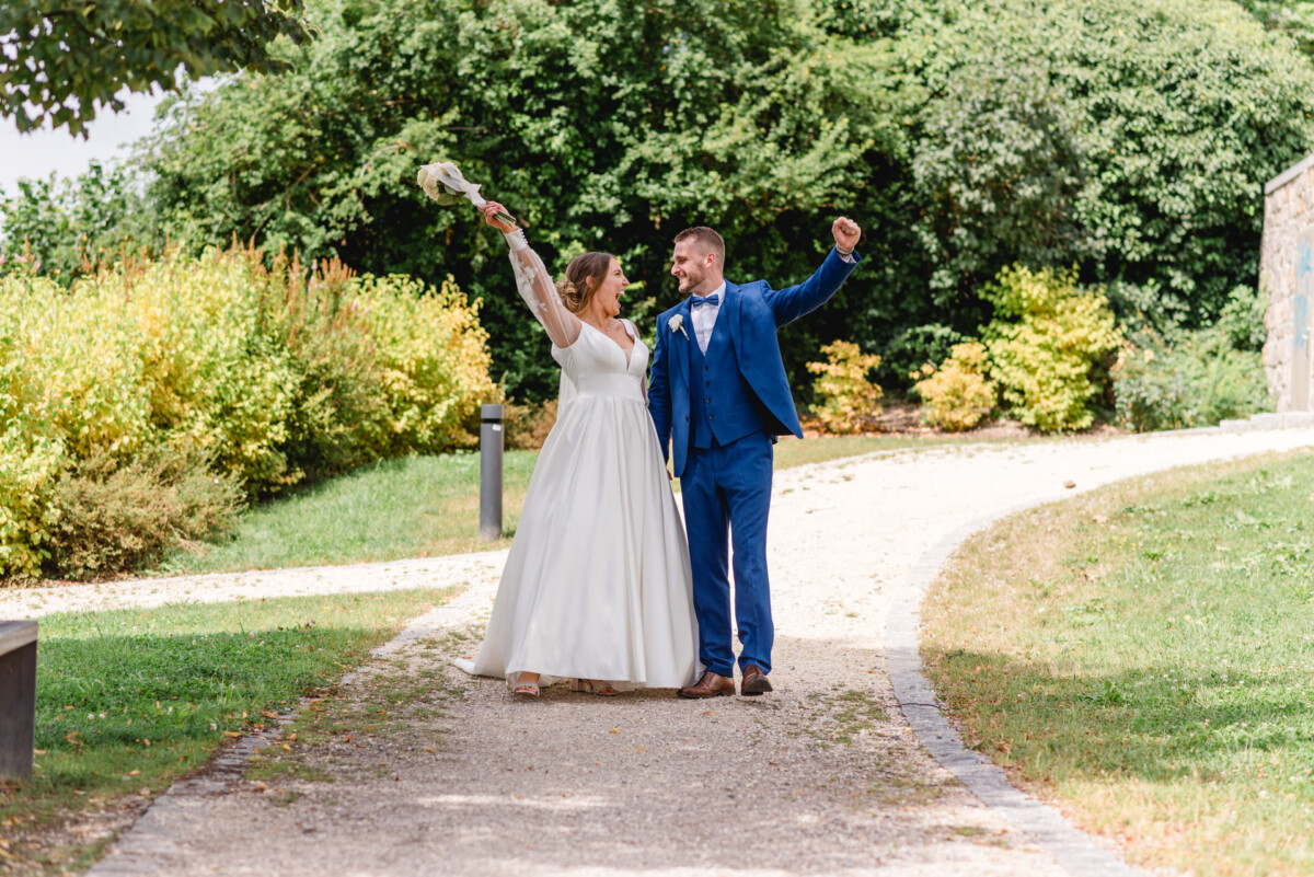 Eine Braut im weißen Kleid und ein Bräutigam im blauen Anzug gehen freudig auf einem Weg in einem Park spazieren, umgeben von grünem Laub.