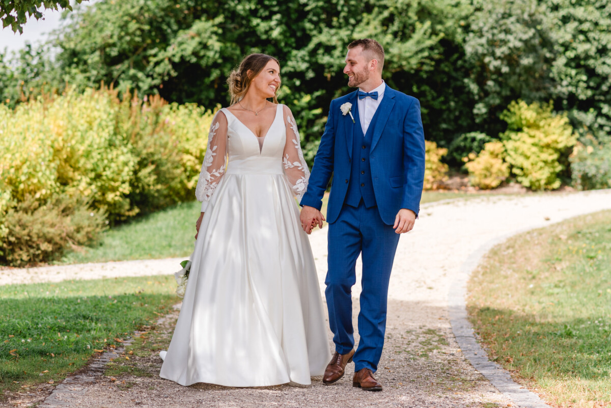 Eine Braut im weißen Kleid und ein Bräutigam im blauen Anzug gehen Hand in Hand einen Gartenweg entlang.