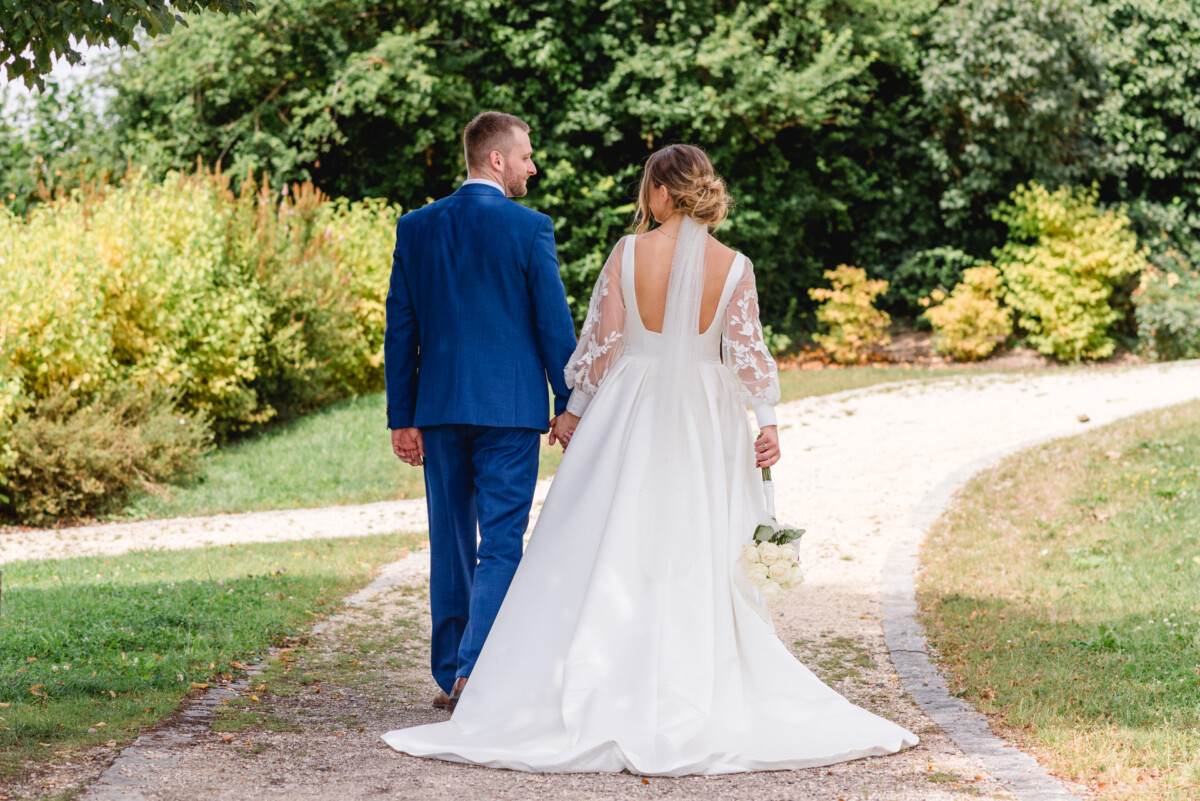 Braut und Bräutigam gehen Hand in Hand einen Gartenweg entlang. Sie trägt ein weißes Kleid mit langer Schleppe, er einen blauen Anzug. Sie sind von Grün umgeben.