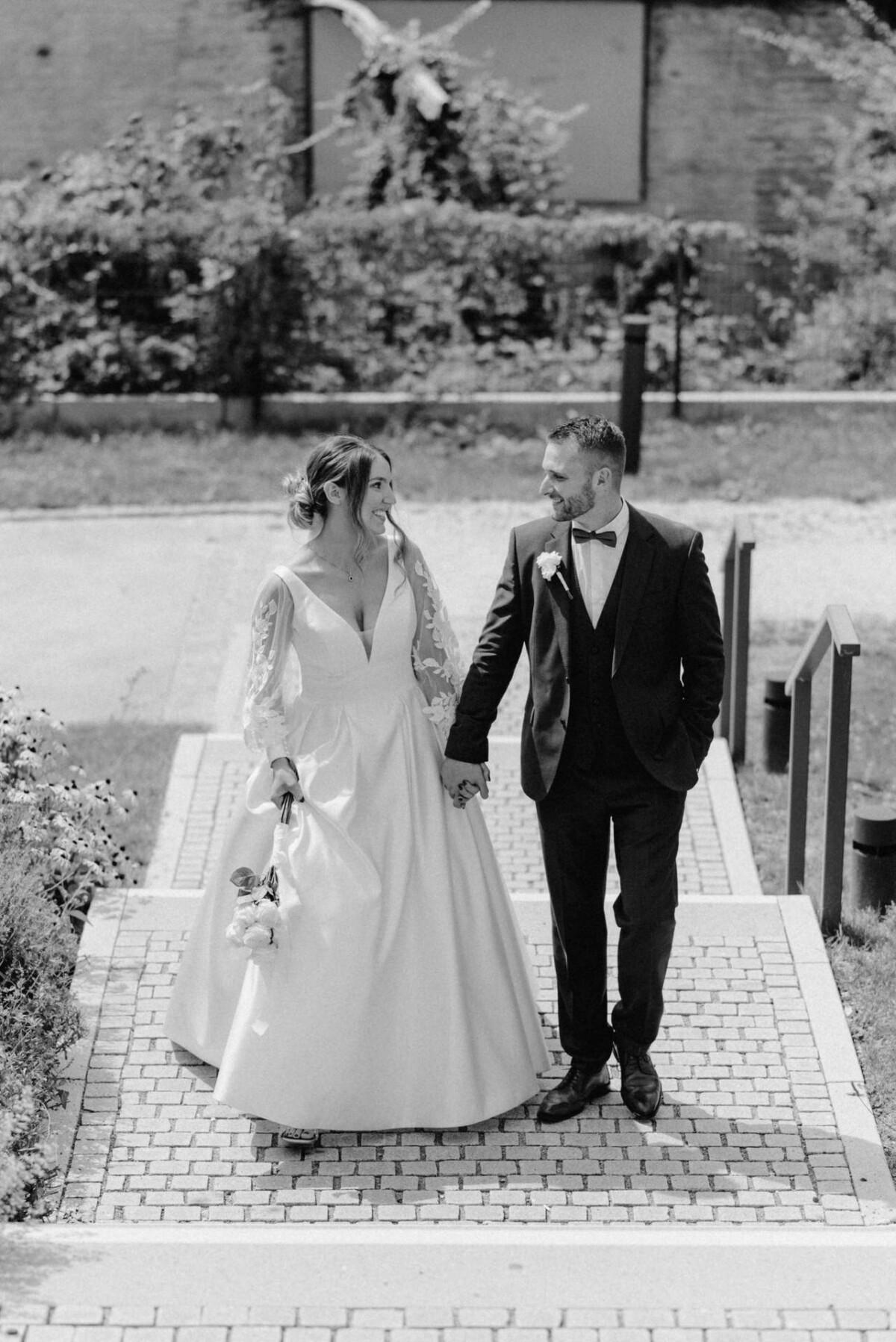 Ein Paar in Abendgarderobe geht Hand in Hand eine Außentreppe hinunter. Die Frau trägt ein langes Kleid, der Mann einen Anzug und eine Fliege. Schwarzweißfoto.