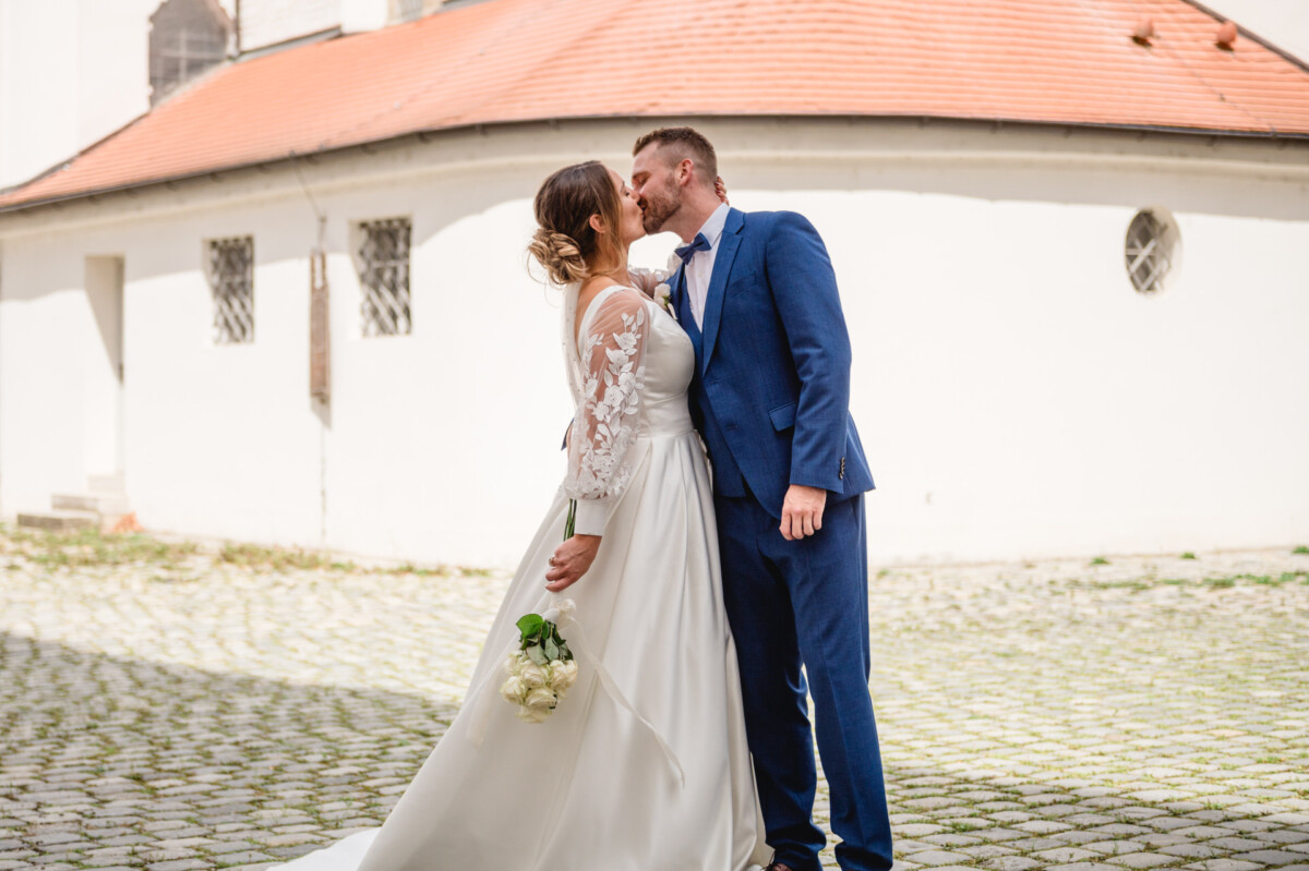 Braut und Bräutigam küssen sich vor einem weißen Gebäude mit rotem Dach. Die Braut hält einen Strauß weißer Blumen.
