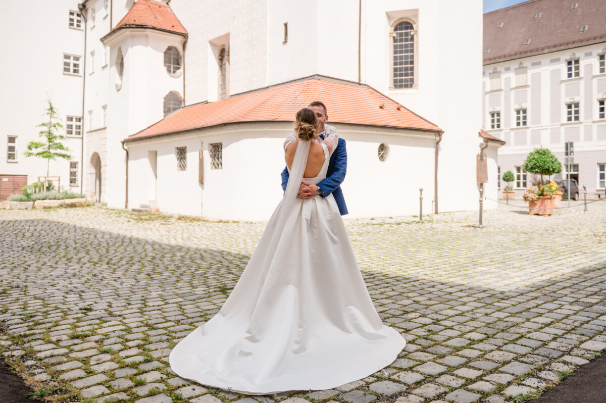 Braut und Bräutigam umarmen sich in einem Innenhof mit historischen Gebäuden im Hintergrund. Die Braut trägt ein langes, weißes Kleid und der Bräutigam einen blauen Anzug.