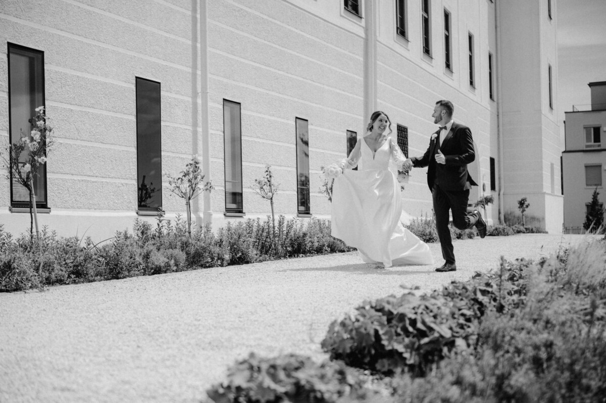 Ein Paar im Hochzeitskleid geht Hand in Hand einen Weg neben einem großen Gebäude entlang, umgeben von Grün.