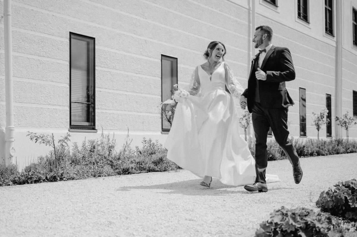 Braut und Bräutigam gehen lächelnd vor einem Gebäude spazieren. Die Braut hält den Saum ihres Kleides, während der Bräutigam Anzug und Fliege trägt. Schwarzweißfoto.