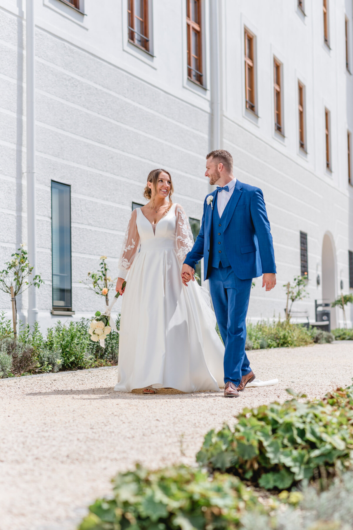 Ein Paar im Hochzeitskleid geht Hand in Hand auf einem Kiesweg neben einem weißen Gebäude mit mehreren Fenstern.