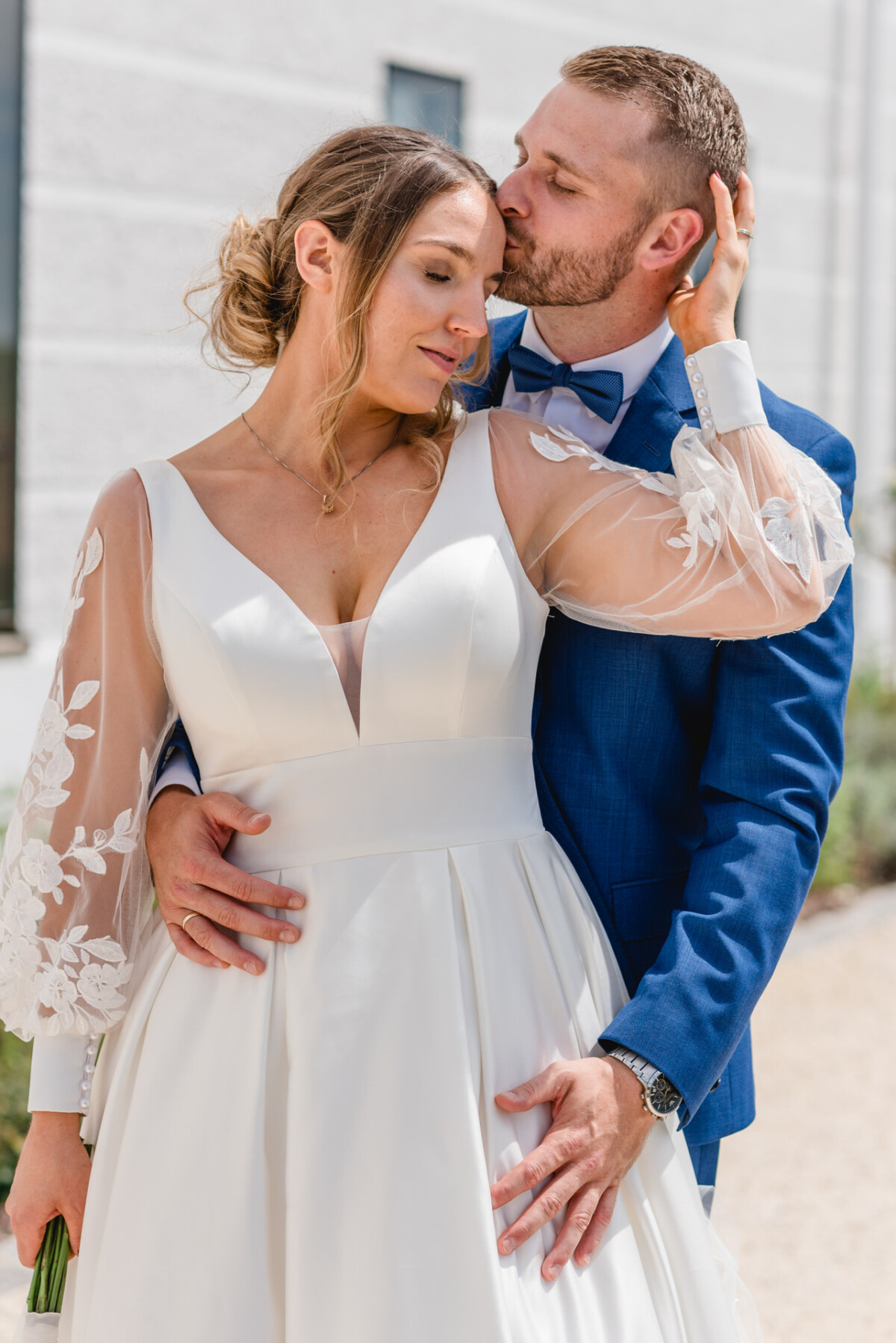 Ein Paar im Hochzeitskleid teilt einen zärtlichen Moment im Freien. Die Braut trägt ein weißes Kleid mit Spitzenärmeln und der Bräutigam einen blauen Anzug mit Fliege.