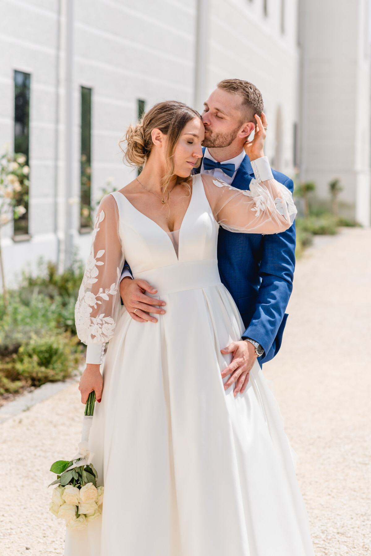 Braut und Bräutigam im Freien, wobei der Bräutigam in einem blauen Anzug den Kopf der Braut küsst. Die Braut hält einen Strauß weißer Blumen und beide stehen vor einem weißen Gebäude.
