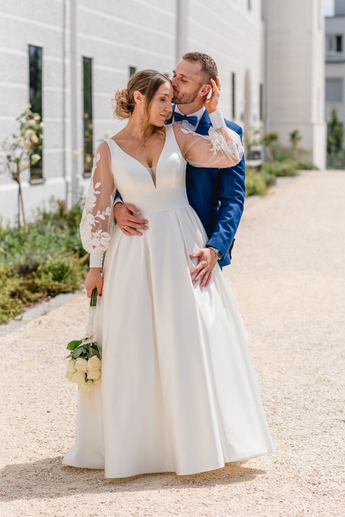 Ein Paar im Hochzeitskleid steht draußen. Die Frau hält einen Blumenstrauß und trägt ein weißes Kleid mit geblümten Ärmeln; der Mann trägt einen blauen Anzug und umarmt sie sanft.