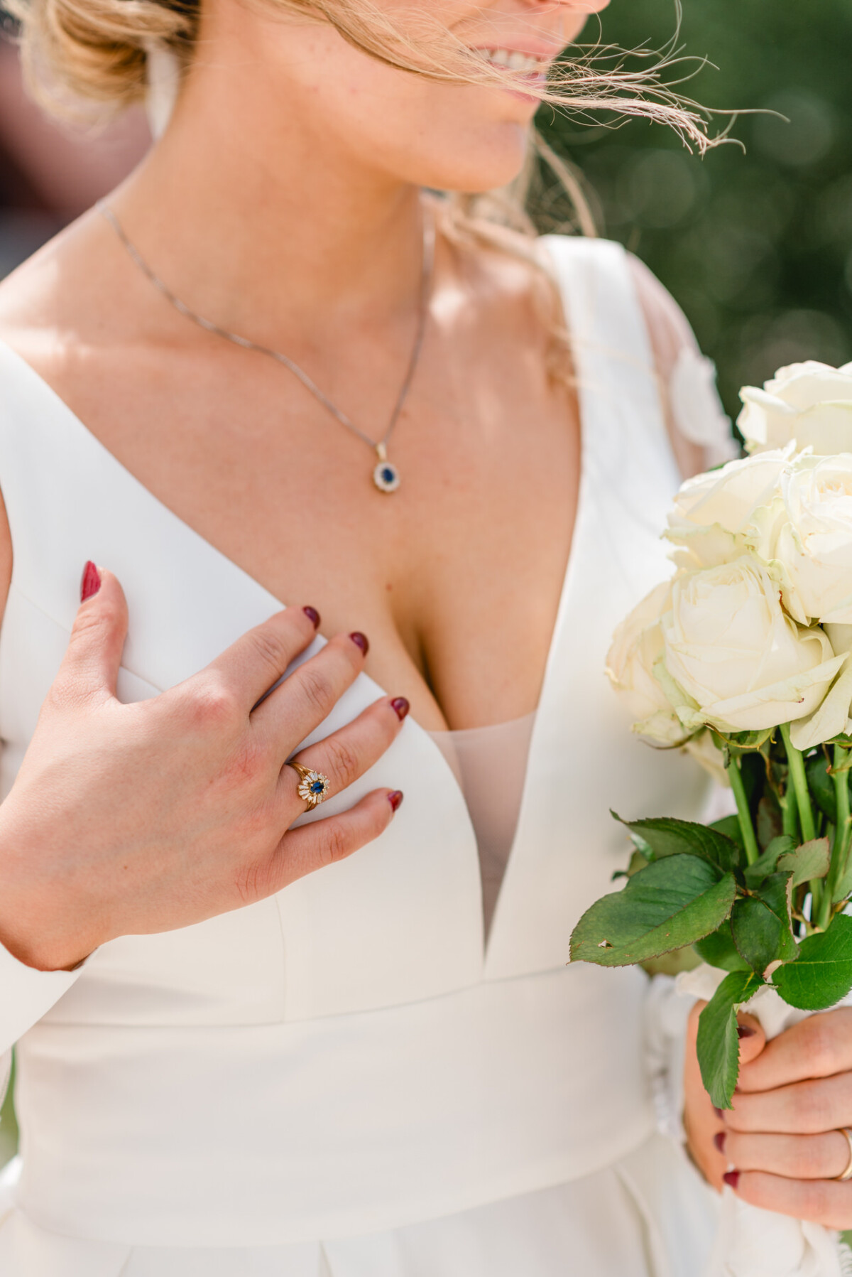 Eine Person im weißen Kleid hält weiße Rosen und präsentiert einen Ring mit einem detaillierten Design. Haare und Schmuck ergänzen das Outfit.