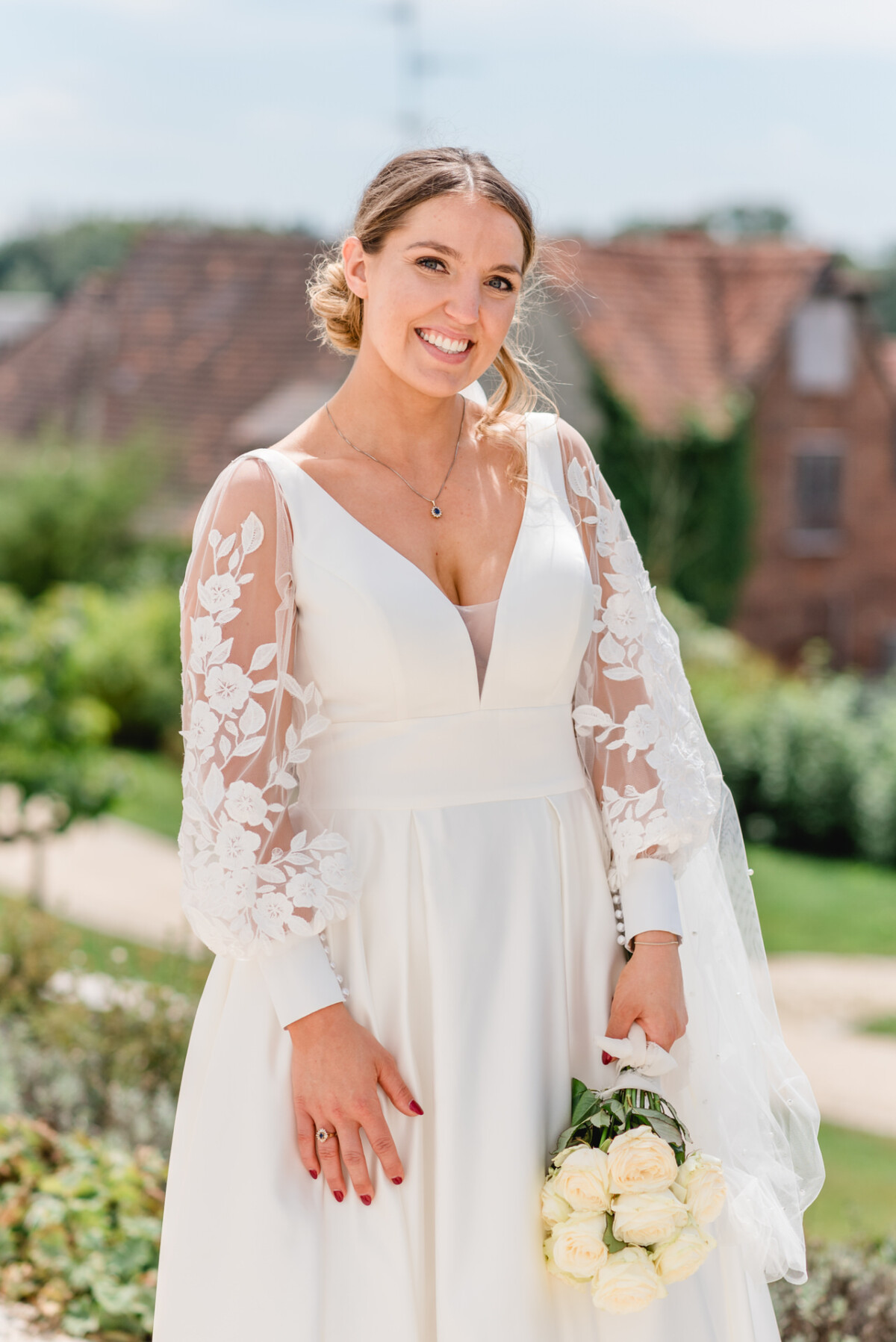 Eine Frau in einem weißen Brautkleid mit Spitzenärmeln hält an einem sonnigen Tag im Freien einen Strauß weißer Rosen.