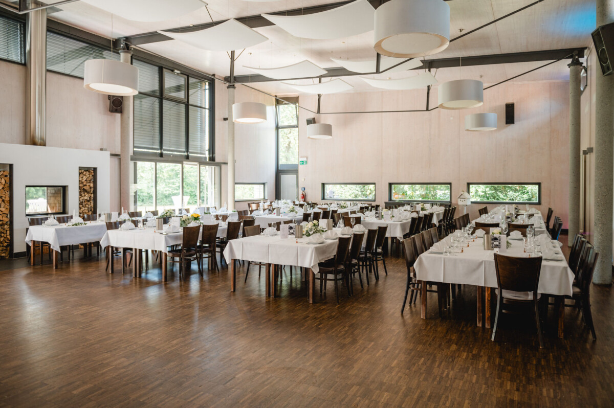 Ein geräumiger Bankettsaal mit mehreren gedeckten Tischen, weißen Tischdecken, Holzstühlen und modernem Deckendekor. Große Fenster lassen den Raum mit natürlichem Licht durchfluten.