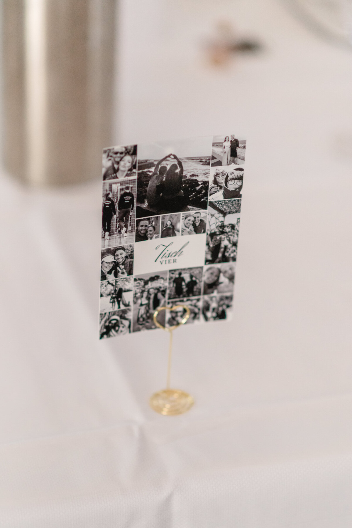 Ein Tischkartenhalter, der eine schwarzweiße Fotocollage mit verschiedenen Personen und dem zentralen Text „Tisch Vier“ auf einem weiß tuchgedeckten Tisch zeigt.