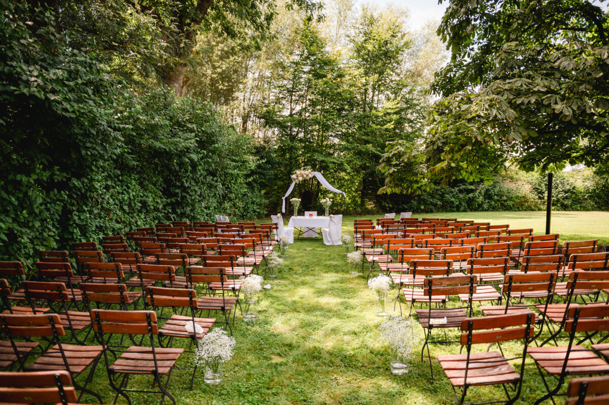 Eine Hochzeitszeremonie im Freien mit Reihen von Holzstühlen vor einem mit weißem Stoff und Blumen geschmückten Altar, umgeben von üppigem Grün.
