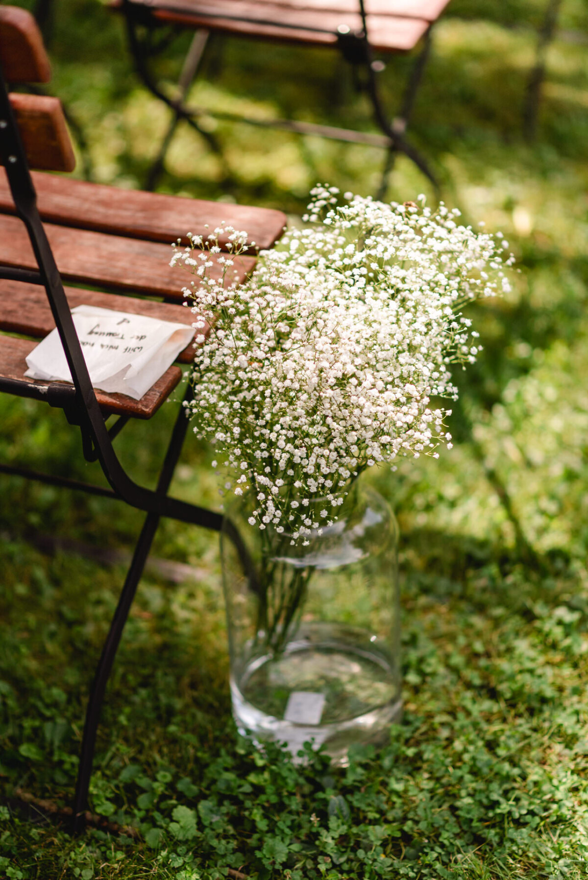 Eine klare Glasvase mit weißen Schleierkrautblüten steht auf einer Wiese neben einem Klappstuhl aus Holz. Auf dem Stuhl liegt ein beschriebenes Blatt Papier.