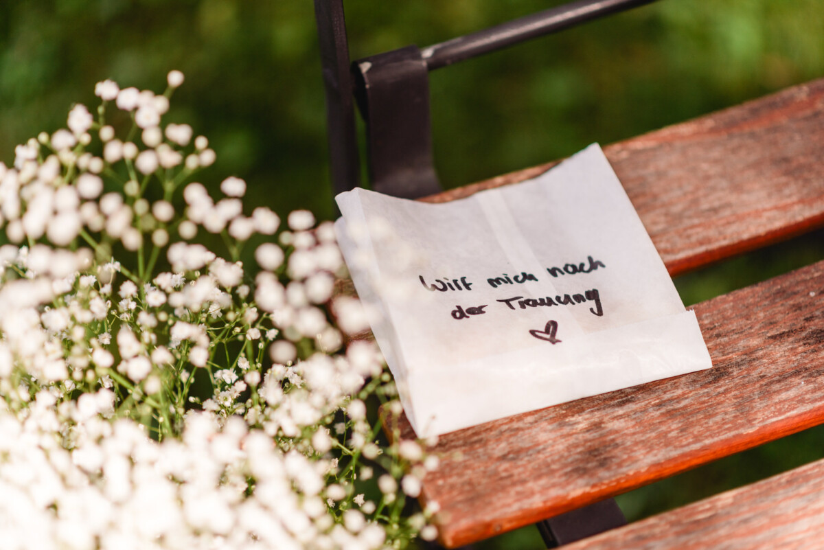 Eine handgeschriebene Notiz in deutscher Sprache auf einem gefalteten Papier mit einem Herzsymbol liegt auf einer Holzbank neben kleinen weißen Blumen.