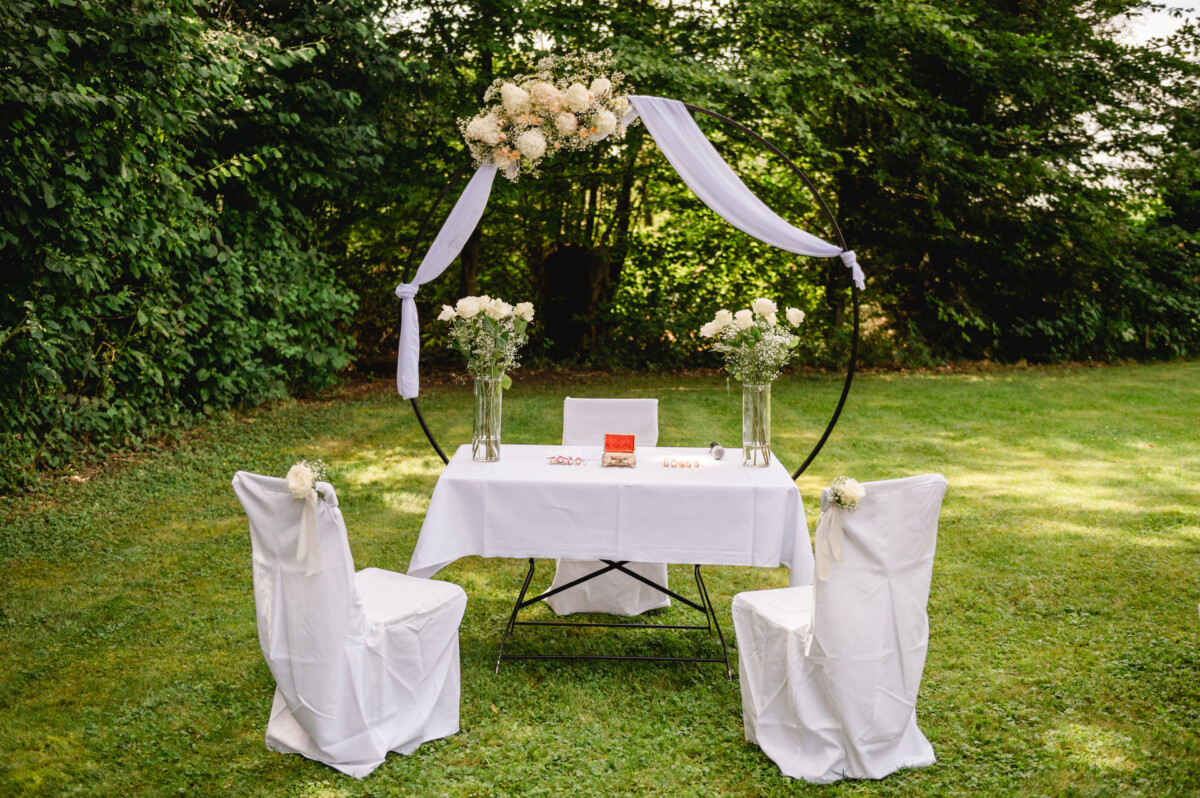 Tischdekoration im Freien für eine Hochzeitszeremonie mit weißen Stühlen, Blumendekorationen und einem Rundbogen.
