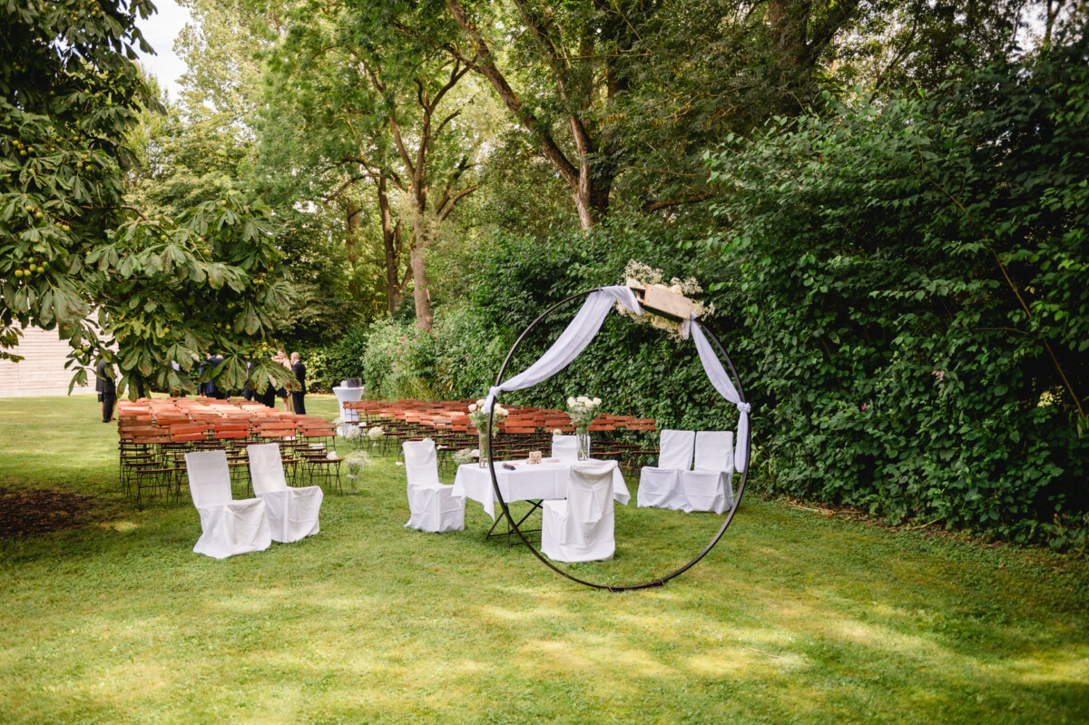 Hochzeitsaufbau im Freien mit einem runden, mit weißem Stoff behangenen Bogen, umgeben von Stühlen und Grünpflanzen.