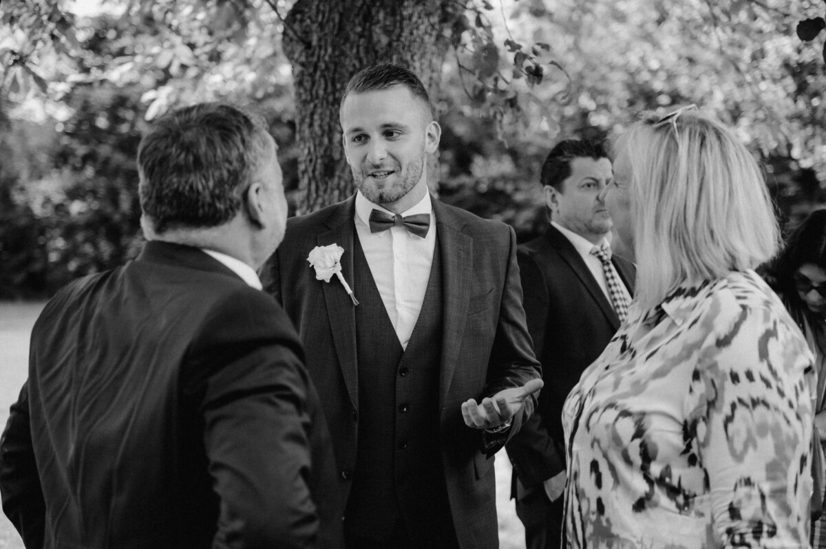 Ein Mann im Anzug mit Fliege unterhält sich im Freien mit zwei Personen. Weitere Personen stehen hinter ihnen, im Hintergrund sind Bäume zu sehen.