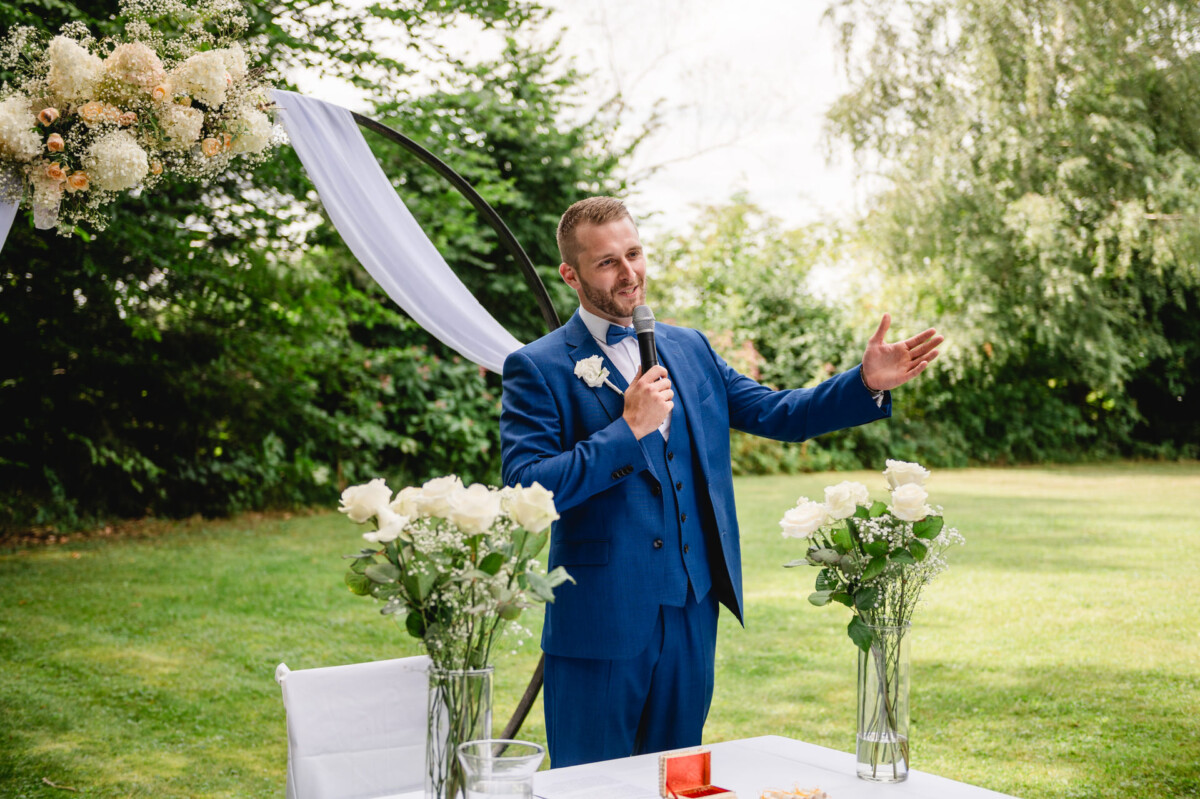 Ein Mann im blauen Anzug hält im Freien eine Rede. Er steht an einem gedeckten Tisch mit zwei Vasen voller weißer Rosen.