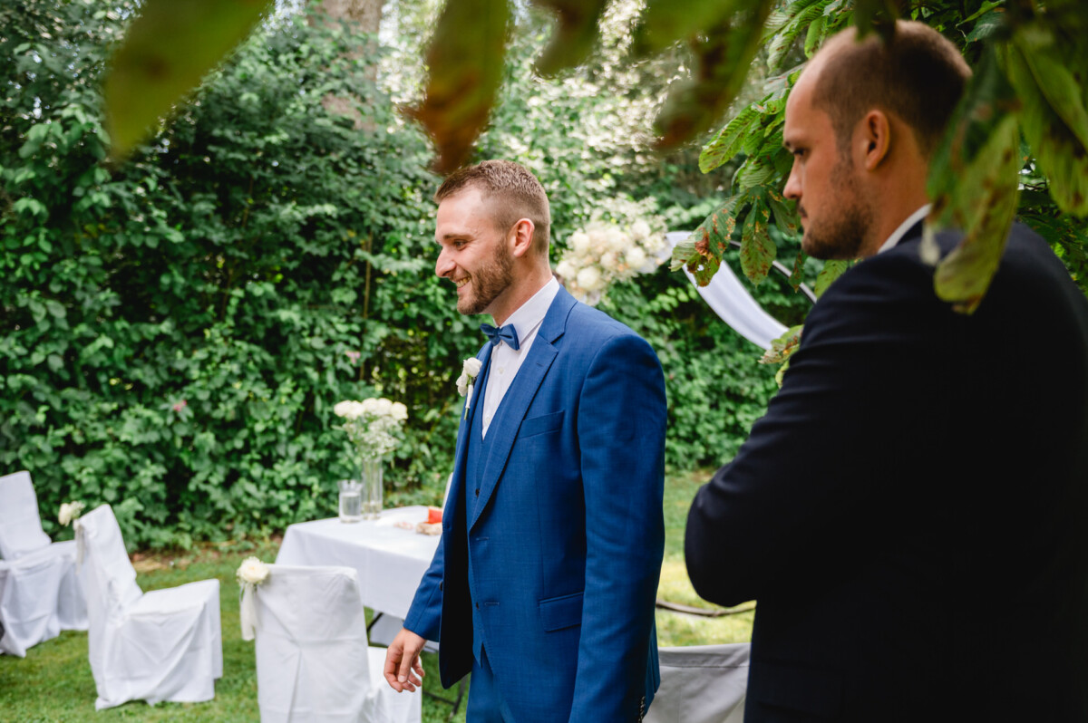 Zwei Männer in Anzügen stehen in einer Außenumgebung mit Stühlen und einem Tisch mit Blumen. Ein Mann trägt einen blauen Anzug und eine Fliege, der andere einen schwarzen Anzug. Der Bereich ist von Grün umgeben.
