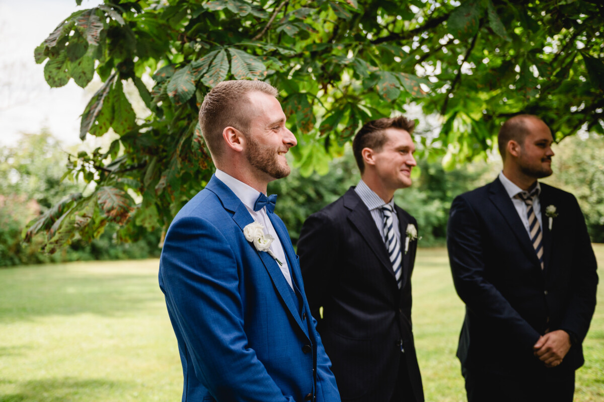 Drei Männer in Anzügen stehen draußen unter einem Baum. Der Mann links trägt einen blauen Anzug. Sie scheinen an einer offiziellen Veranstaltung teilzunehmen.