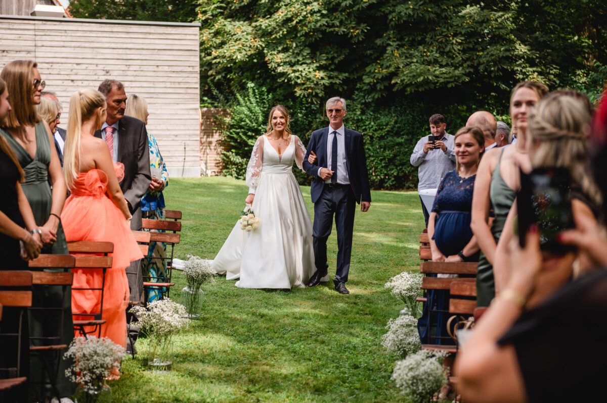 Eine Braut in einem weißen Kleid schreitet mit einem älteren Mann, umgeben von sitzenden Gästen, einen Gang im Freien entlang.