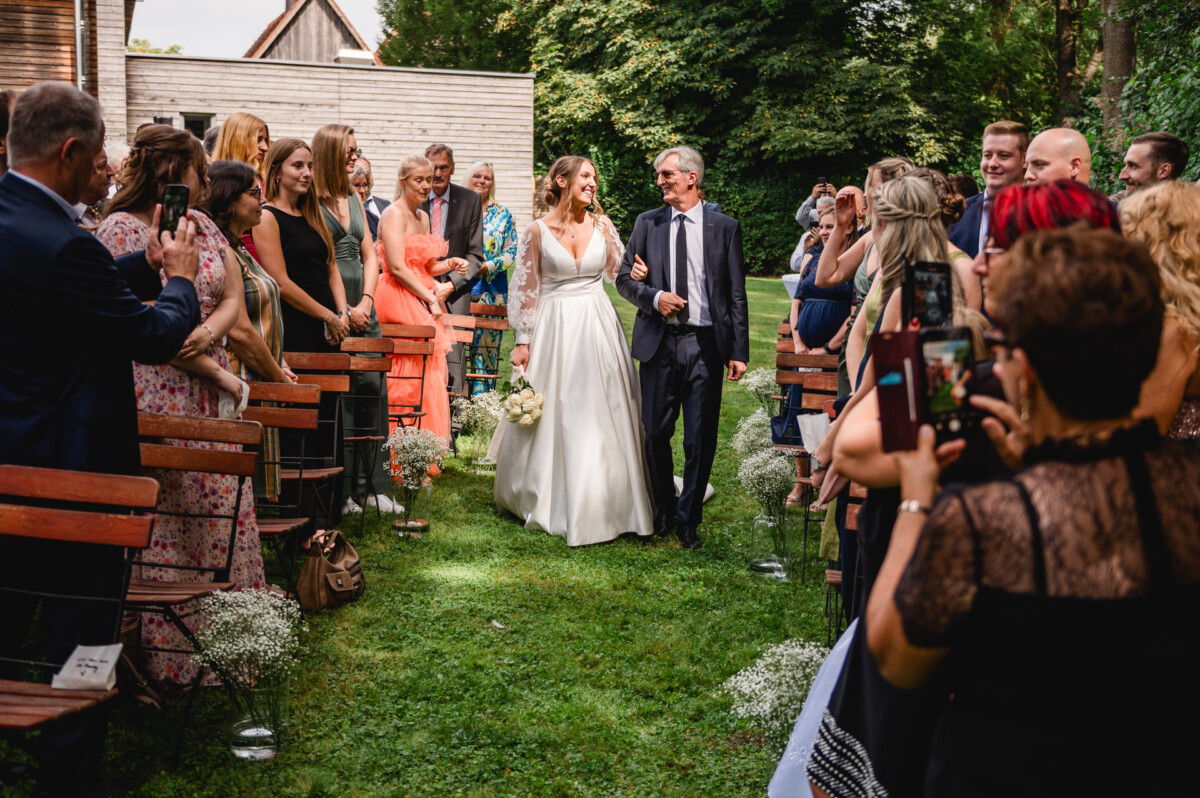 Eine Braut im weißen Kleid schreitet in Begleitung eines Mannes im Anzug und umgeben von stehenden Gästen einen Gang im Freien entlang.