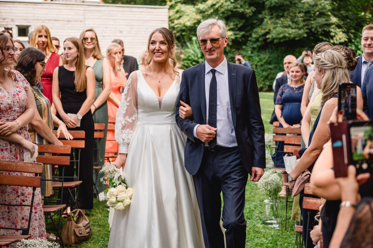Eine Braut im weißen Kleid schreitet mit einem älteren Mann im Anzug einen Gang im Freien entlang, umgeben von sitzenden und stehenden Gästen.