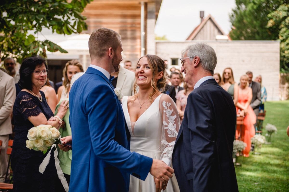 Eine Braut in einem weißen Kleid steht mit einem Bräutigam und einem älteren Mann in Anzügen, umgeben von Hochzeitsgästen, vor der Tür.