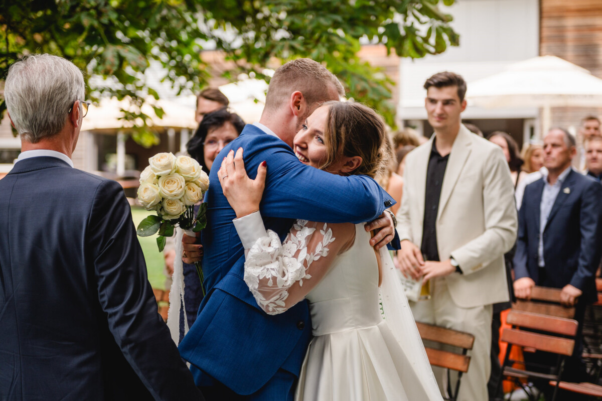 Eine Braut im weißen Kleid umarmt einen Mann im blauen Anzug und hält dabei einen Strauß weißer Rosen in der Hand. Umgeben sind sie von Hochzeitsgästen.