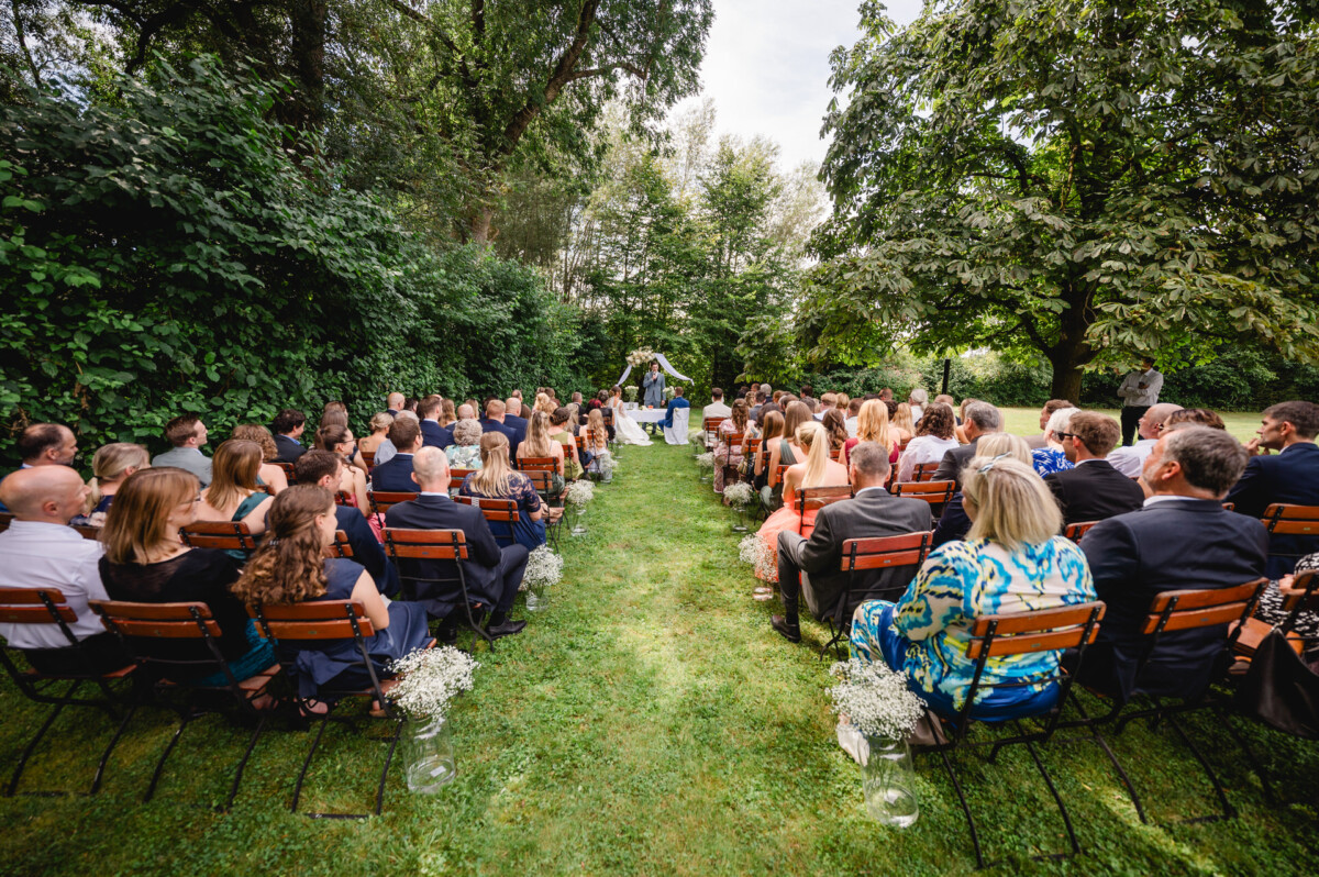 Hochzeitszeremonie im Freien mit Gästen, die auf beiden Seiten eines Ganges sitzen. Ein Paar steht unter einem Baumdach am Altar.
