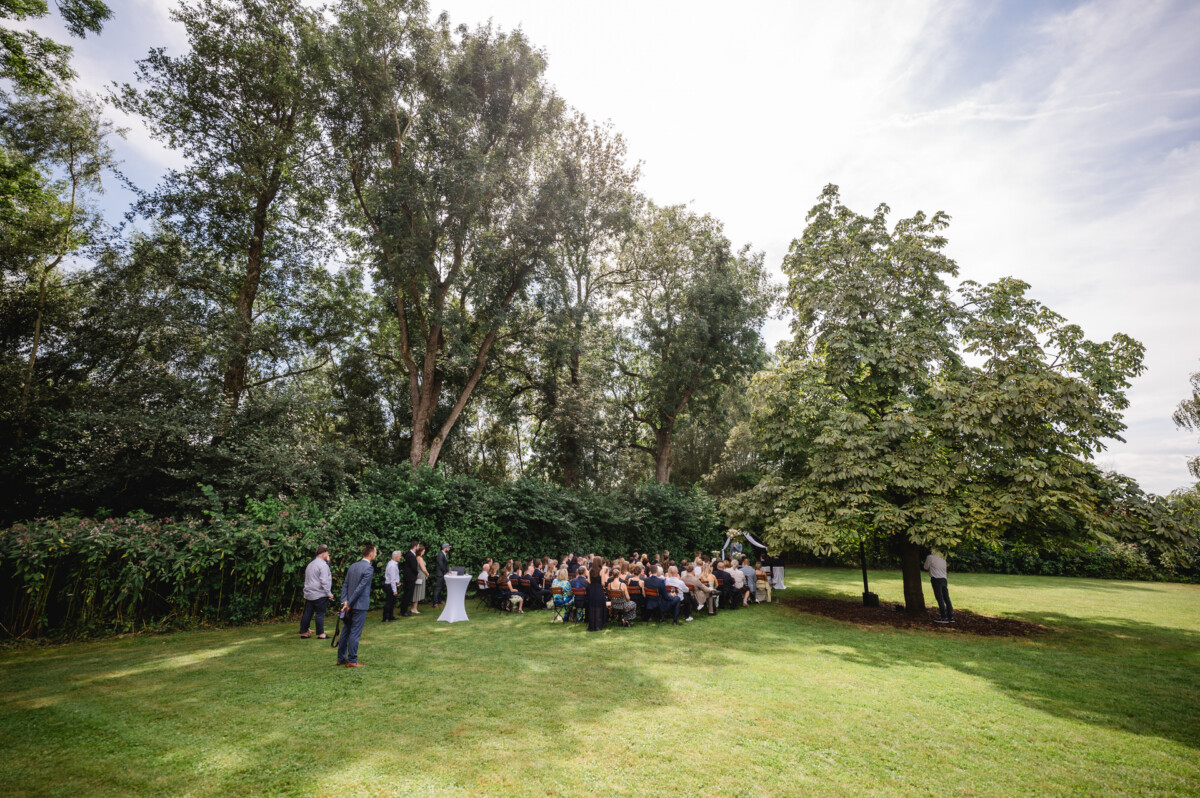 Eine Gruppe von Menschen hat sich in einer grasbewachsenen Umgebung im Freien versammelt, umgeben von Bäumen und Grün; einige sitzen, andere stehen.