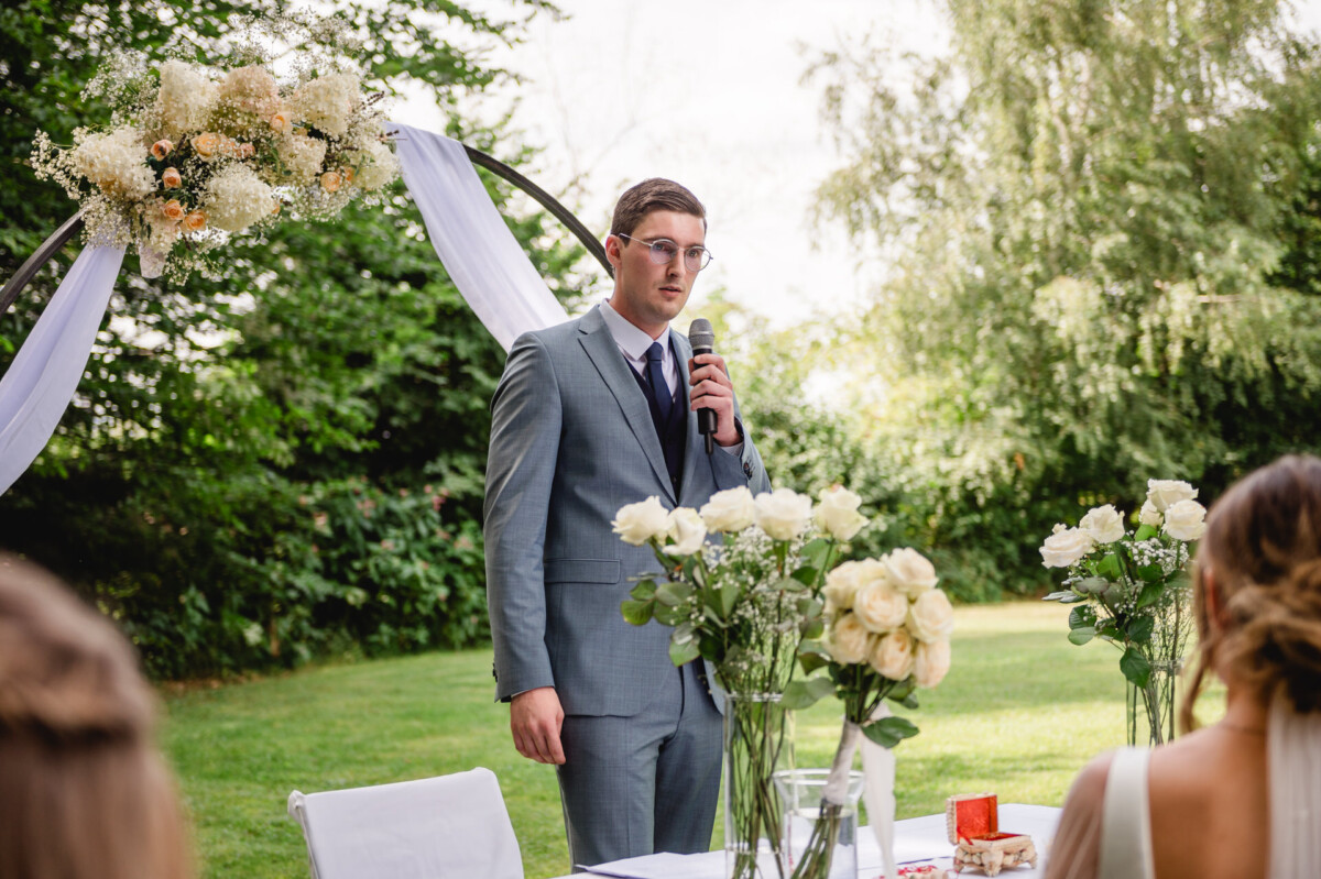 Eine Person in einem grauen Anzug spricht im Freien in ein Mikrofon, in der Nähe eines Blumenbogens und weißer Rosen auf Tischen.