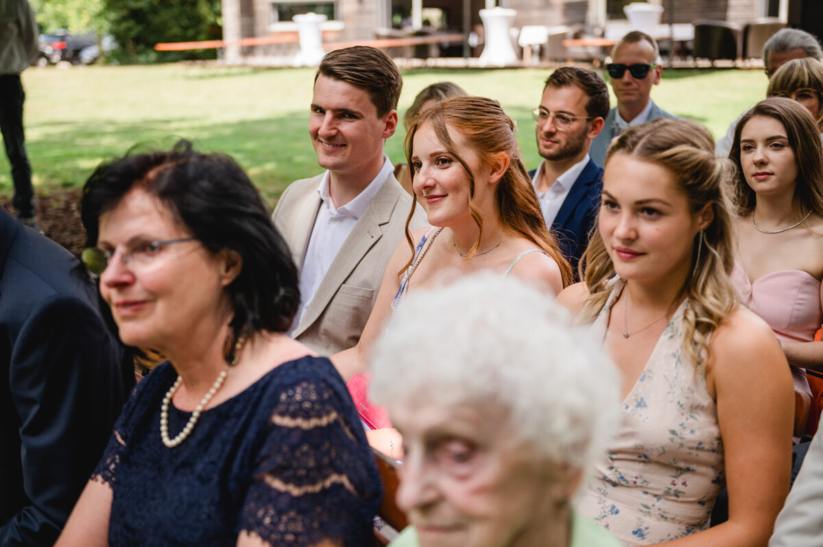 Eine Gruppe von Menschen sitzt draußen auf einer Rasenfläche und nimmt an einer Veranstaltung teil. Einige sind formell gekleidet und alle blicken nach vorne.