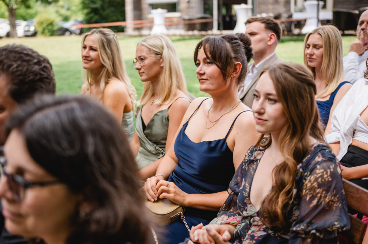 Eine Gruppe von Menschen sitzt im Freien und beobachtet aufmerksam ein Ereignis. Einige sind in formelle Kleidung gekleidet und von Grün umgeben.