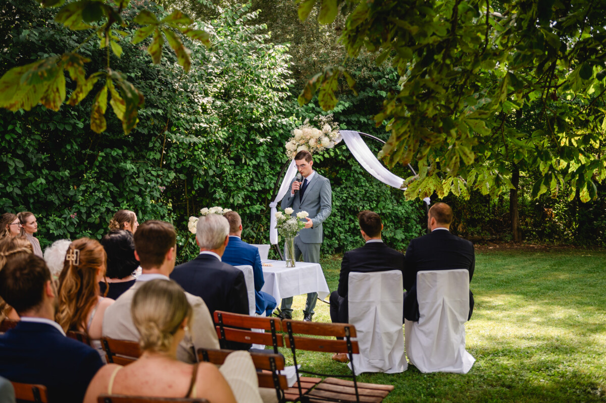 Eine Person im Anzug steht an einem kleinen Tisch und spricht in ein Mikrofon während einer Zeremonie im Freien, an der sitzende Gäste unter belaubten Bäumen teilnehmen.