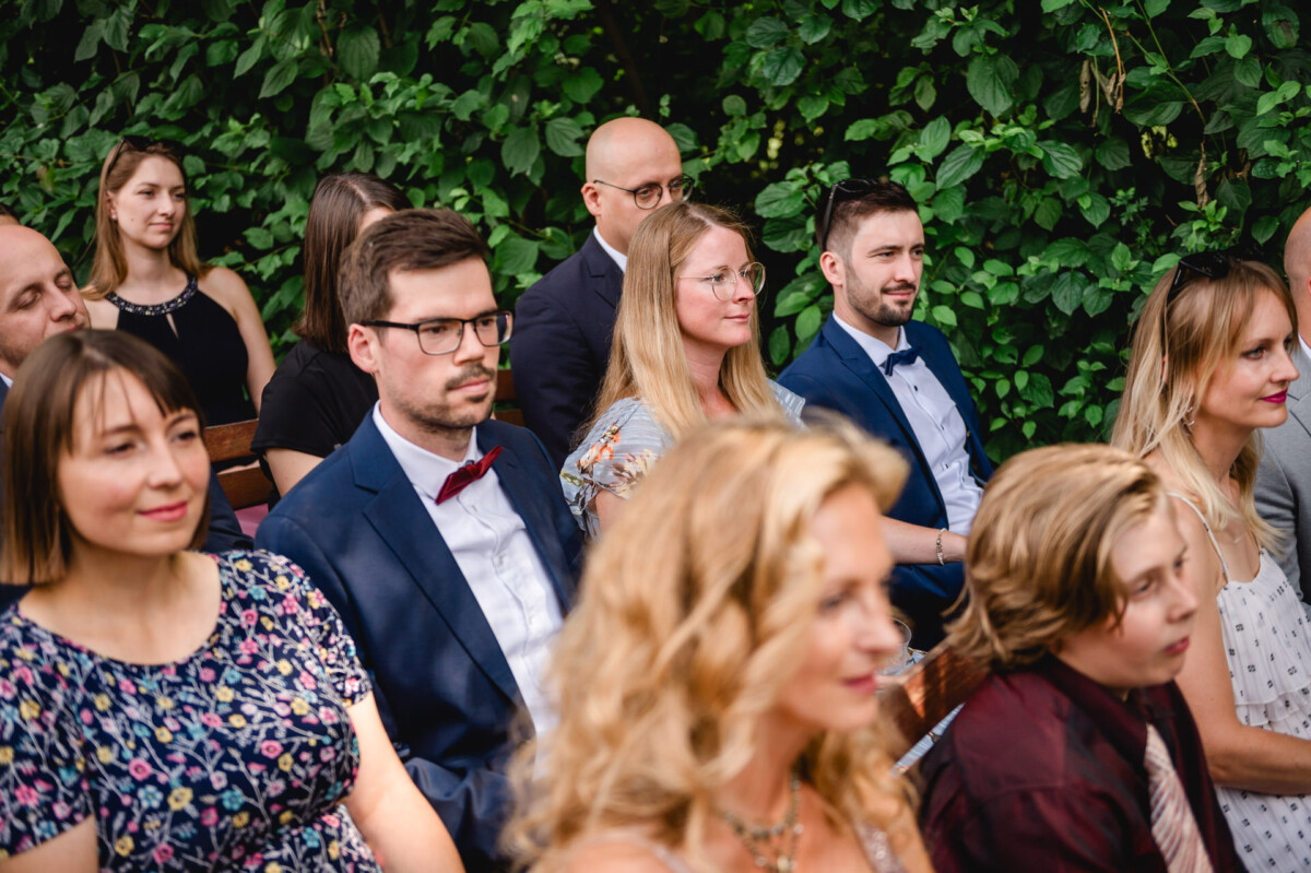 Eine Gruppe von Menschen in Abendgarderobe sitzt im Freien, im Hintergrund grünes Laub.