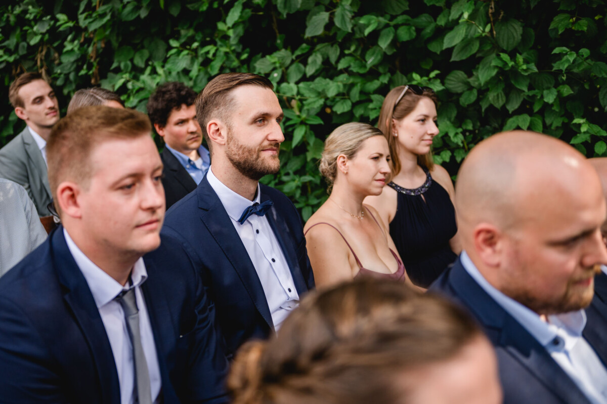 Eine Gruppe von Menschen in formeller Kleidung sitzt im Freien, im Hintergrund grünes Laub. In der Mitte steht ein Mann in Anzug und Fliege.