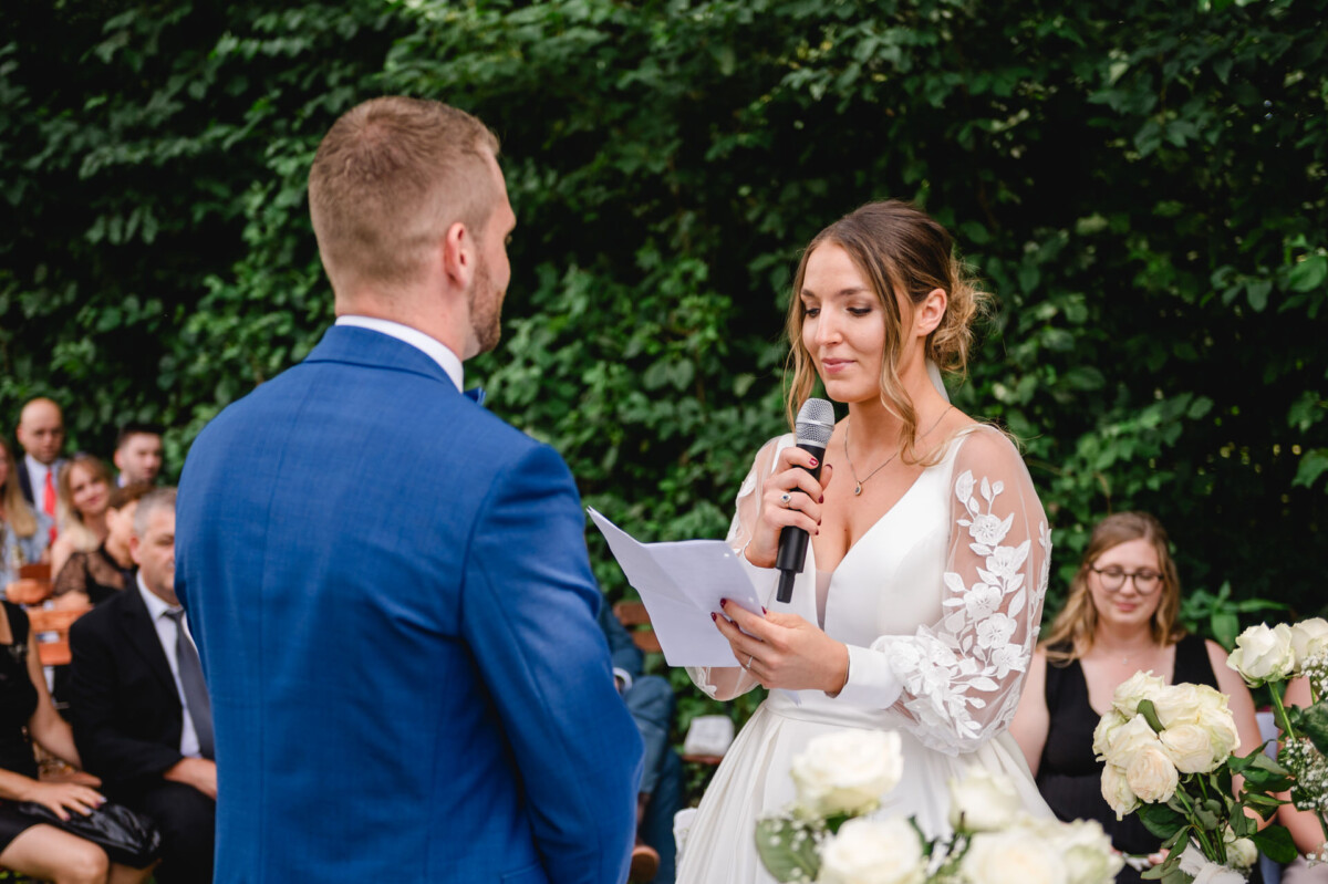 Eine Braut in einem weißen Kleid liest aus einer Zeitung, während sie ein Mikrofon hält, und blickt einem Bräutigam in einem blauen Anzug während einer Zeremonie im Freien gegenüber. Im Hintergrund sitzen Menschen. Weiße Rosen sind zu sehen.