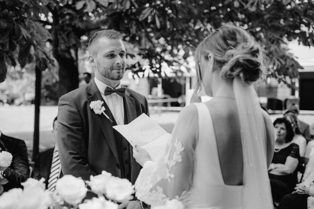 Eine Braut liest dem Bräutigam während einer Hochzeitszeremonie im Freien ihre Ehegelübde vor. Beide sind formell gekleidet, im Hintergrund sitzen die Gäste.