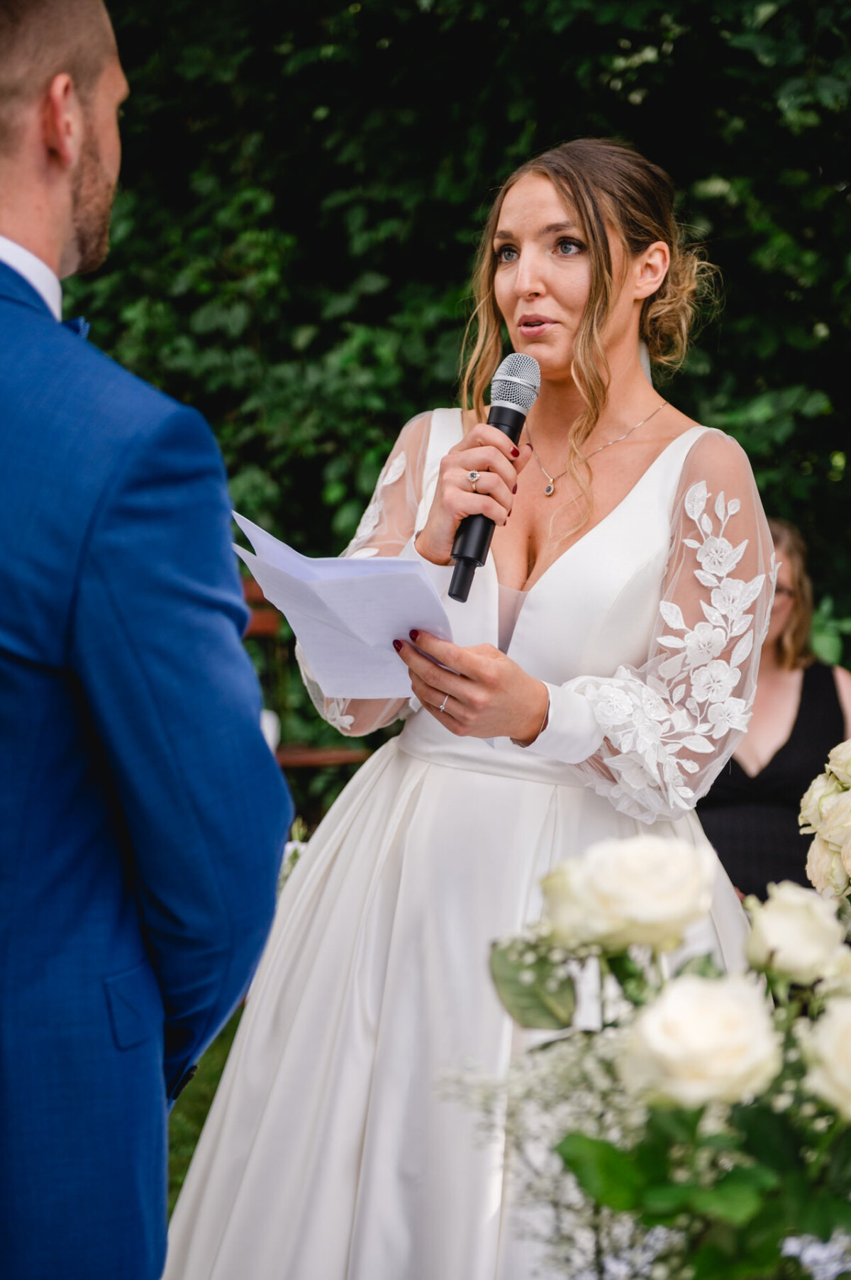 Eine Braut in einem weißen Kleid mit geblümten Ärmeln liest während einer Hochzeitszeremonie im Freien ihre Gelübde in ein Mikrofon. Im Hintergrund sind Blumen und Grünpflanzen zu sehen.