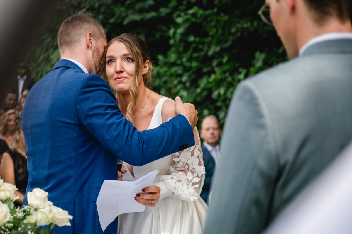 Eine Braut im weißen Kleid umarmt ihren Bräutigam im blauen Anzug während einer Zeremonie im Freien, umgeben von Gästen.