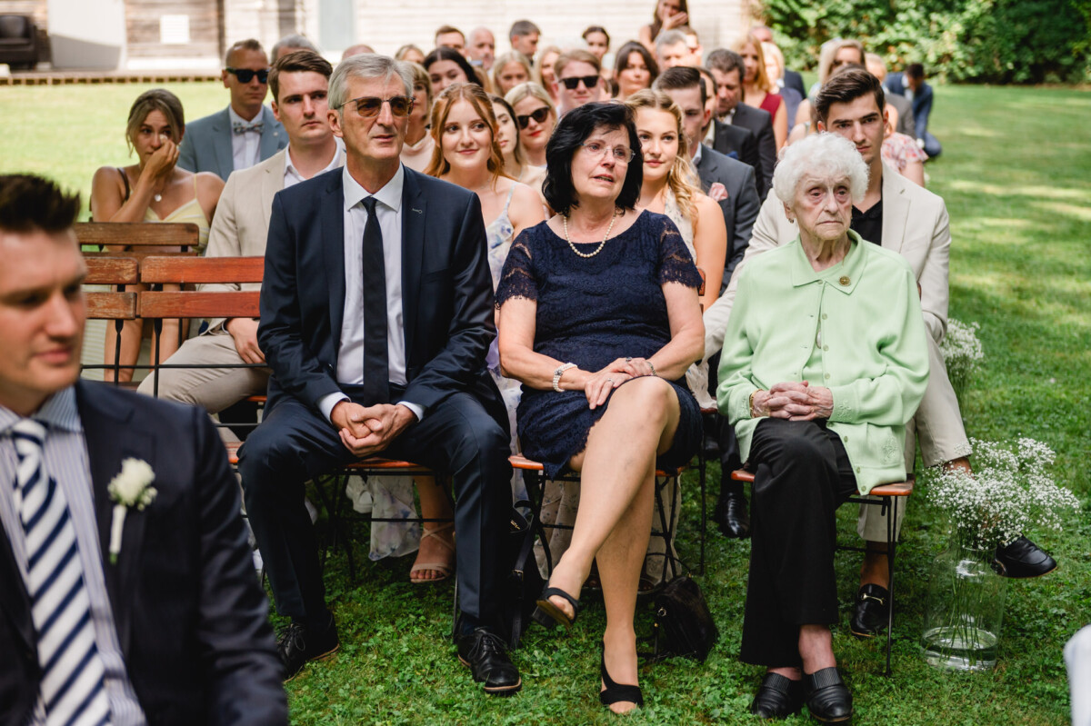 Eine Gruppe von Menschen, formell gekleidet, sitzt im Freien auf Holzstühlen, mit Grünflächen und einem Gebäude im Hintergrund.