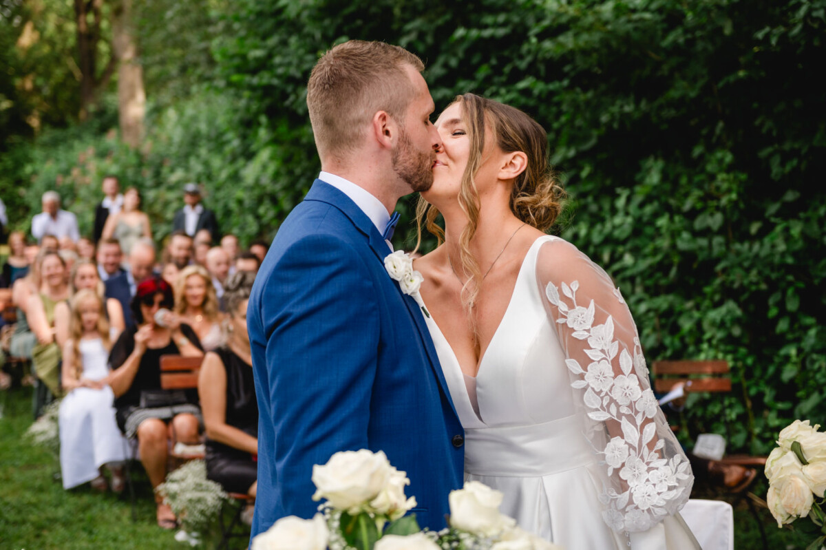 Ein Paar küsst sich bei einer Hochzeit im Freien, umgeben von Gästen, die in einer Gartenkulisse sitzen. Im Vordergrund sind Blumen zu sehen.