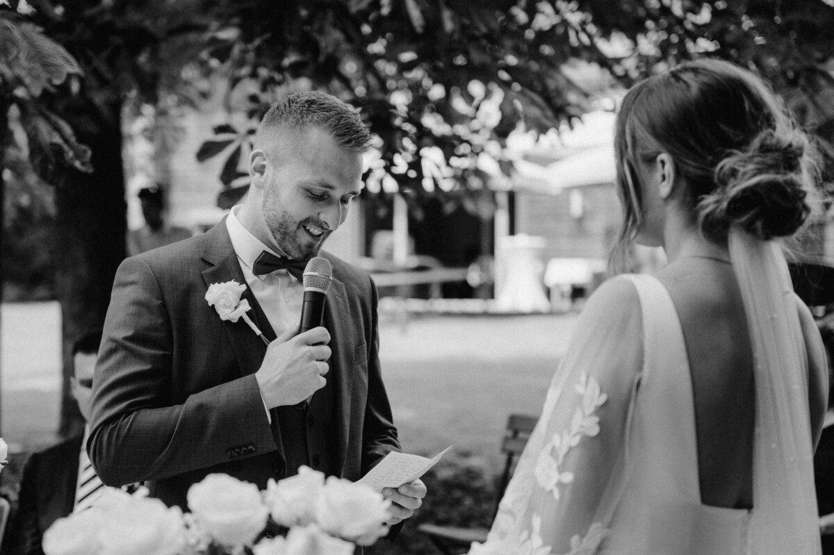 Ein Mann im Anzug liest aus einer Zeitung, während er ein Mikrofon hält. Er steht neben einer Frau im Brautkleid im Freien.