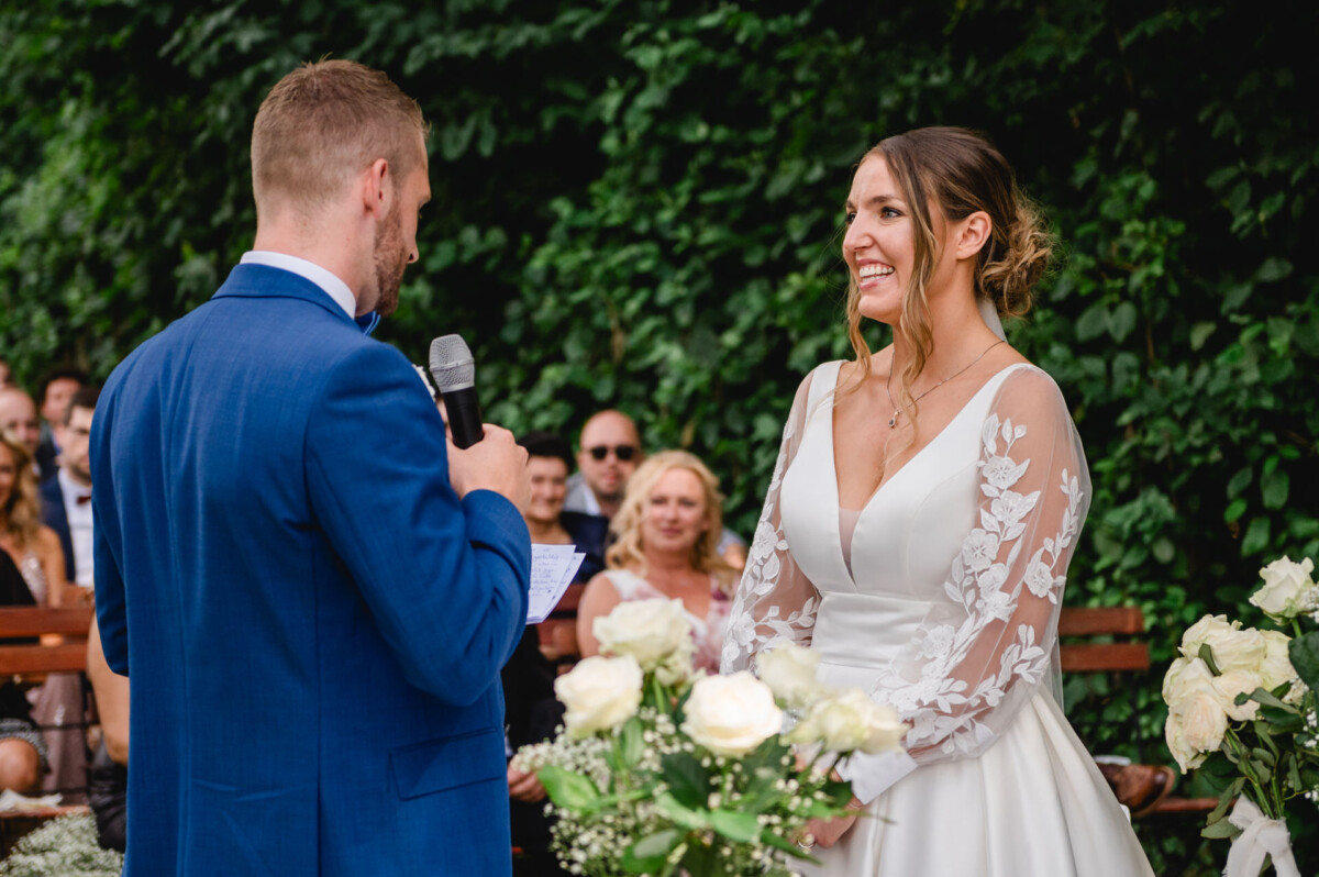 Ein Mann im blauen Anzug spricht während einer Hochzeitszeremonie in ein Mikrofon, während eine Frau im weißen Brautkleid vor dem Publikum lächelt.
