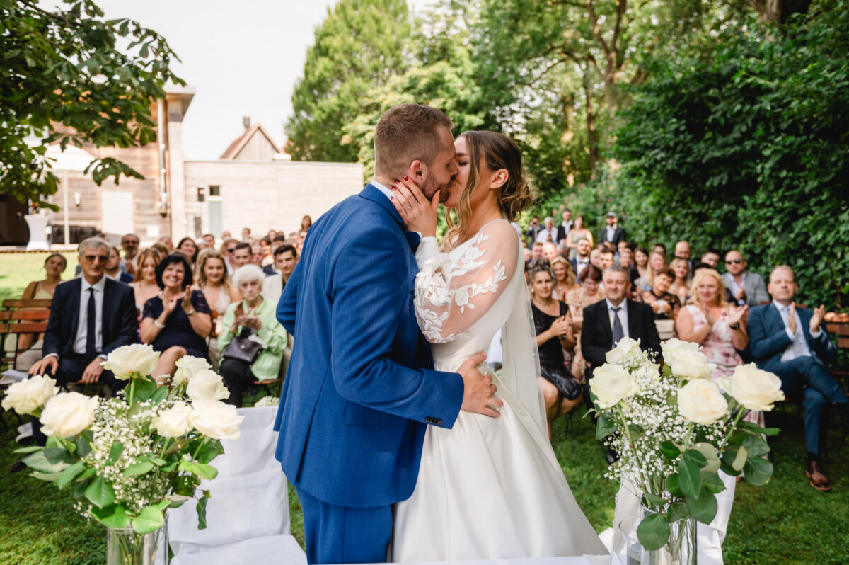 Braut und Bräutigam küssen sich bei einer Hochzeitszeremonie im Freien, umgeben von auf Bänken sitzenden Gästen, im Vordergrund sind weiße Rosen zu sehen.