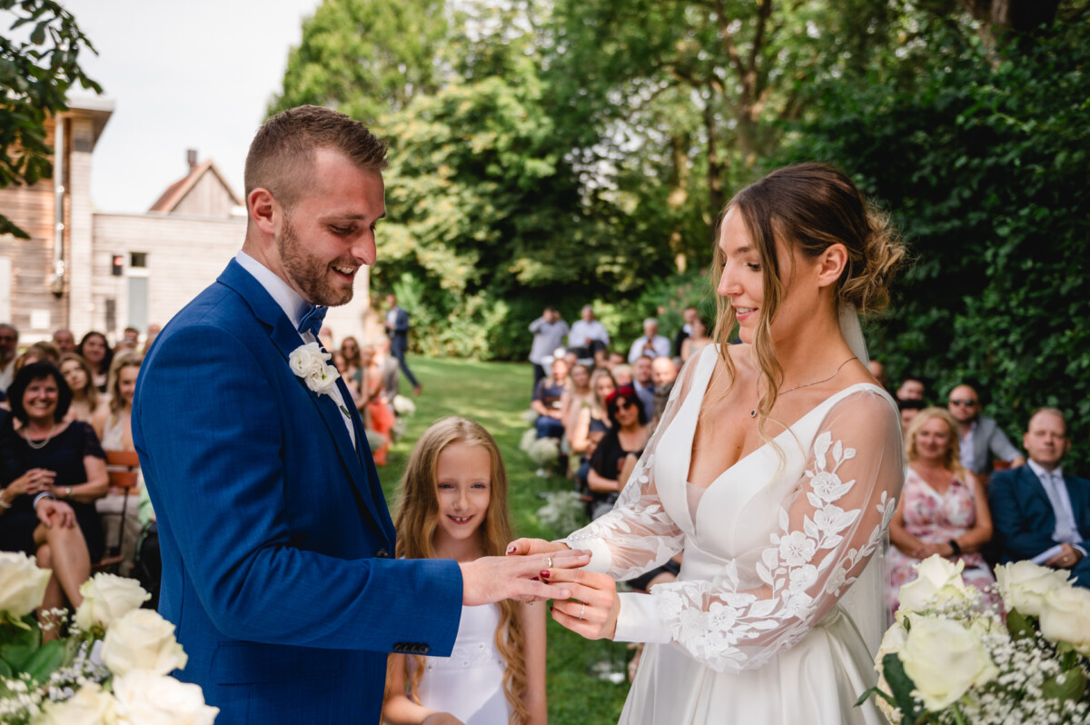 Ein Paar tauscht während einer Hochzeitszeremonie im Freien Ringe aus. Ein junges Mädchen steht in der Nähe und im Hintergrund sitzen Gäste.